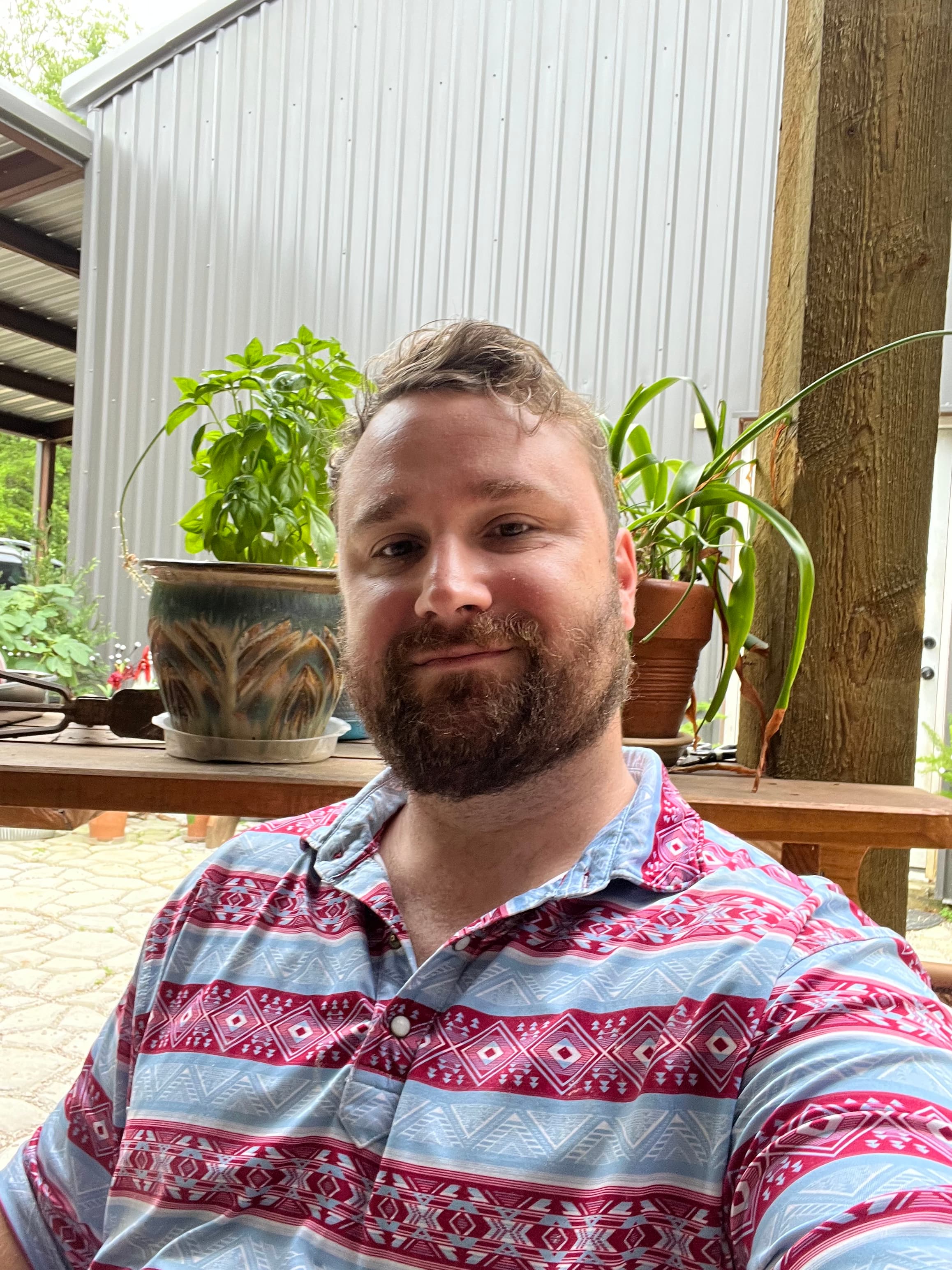 Bearded man wearing a red and blue geometric patterned shirt posing outdoors with potted plants.